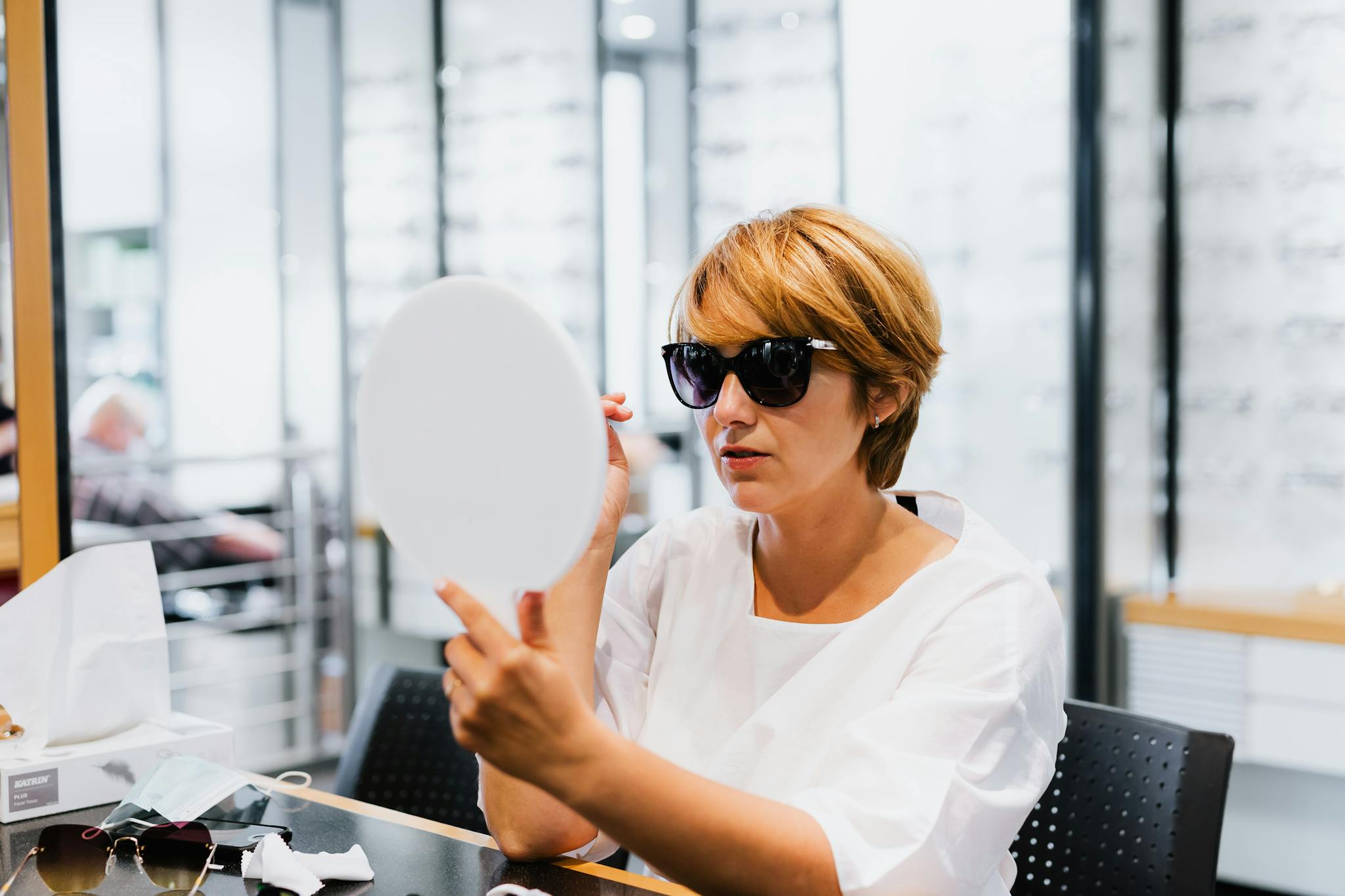 A woman checks her new sunglasses using a hand mirror in an optical store.