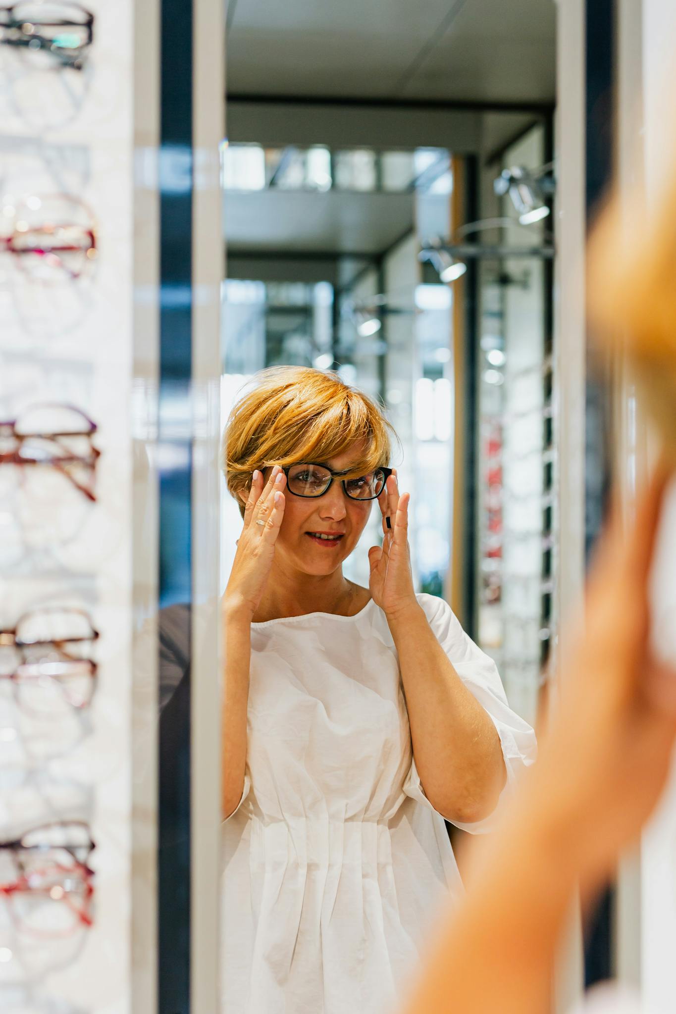 Adult woman trying on eyeglasses at an optical store, looking in a mirror.